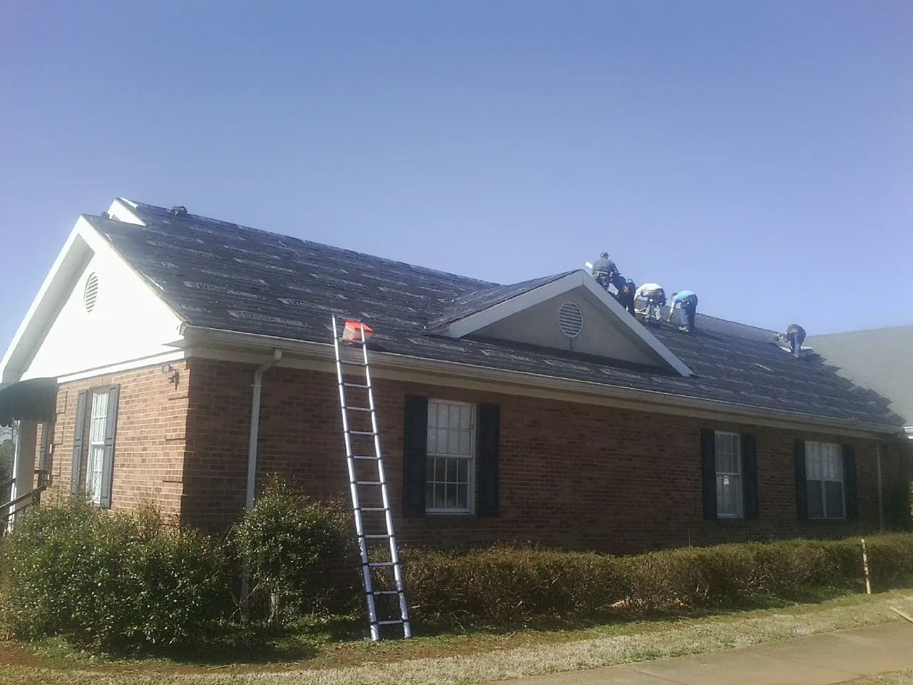 Crew tearing shingles off a brick ranch roof with a ladder set — Emergency Roof Repair work by our crew in Hamer
