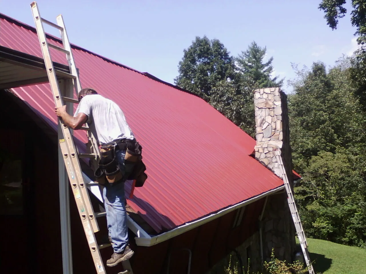 Roofer on a ladder beside a red metal roof and stone chimney — Soffit & Fascia Repair by Roofing Brothers in Hamer
